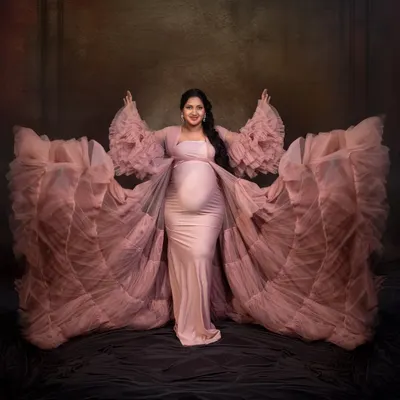Embracing the power and beauty of motherhood. This mom-to-be shows off our popular pink ruffled gown, its dramatic sleeves creating a stunning, angelic silhouette against a classic painterly backdrop.