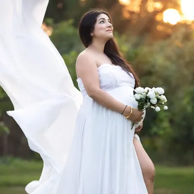 An outdoor maternity portrait taken during the golden hour, with soft sunlight creating a warm, ethereal glow. The flowing white gown and natural setting provide a romantic and serene feel.
