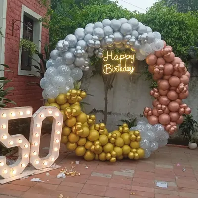 This outdoor setup for a 50th birthday party features a large circular balloon arch in silver, gold, and rose gold. The design is completed with a 'Happy Birthday' neon sign and large marquee numbers, perfect for garden or backyard celebrations.