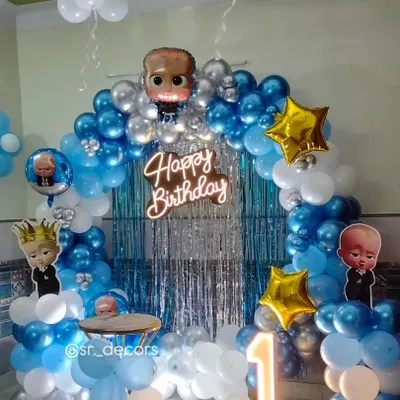 A 'Boss Baby' themed first birthday setup, complete with character cutouts, a blue and silver balloon arch against a shimmer curtain, and a large light-up number '1'.