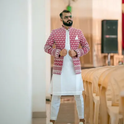 A client looking stylish in an open designer Jodhpuri set. The intricately embroidered red jacket is layered over a simple white kurta and pants, making it a perfect outfit for a Sangeet or wedding celebration.