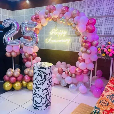 Here is another take on a 25th anniversary, using a soft pink and gold color scheme. A lush balloon arch frames the space, complemented by a 'Happy Anniversary' neon sign and large silver foil balloons announcing the '25' year milestone.