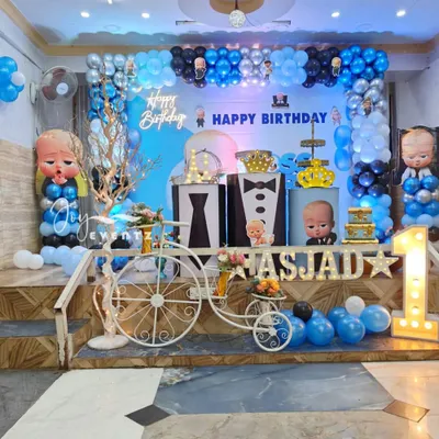 This elaborate Boss Baby stage setup for a first birthday features a multi-level platform, a large balloon arch in shades of blue, black, and silver, and a decorative white bicycle prop. The design also includes character pillars and light-up marquee letters.