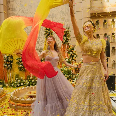 A Haldi ceremony is a festival of joy. Here, the bride and her friend dance with colorful fans amidst a shower of flower petals, capturing the high-energy and vibrant spirit of the event.