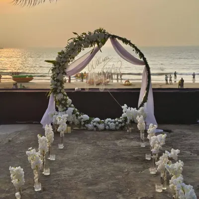 A wide view of a complete beach proposal setup I arranged. The scene includes the main floral arch, a flower-lined aisle with candles in glass vases for a magical pathway, and the stunning backdrop of the sea at sunset.
