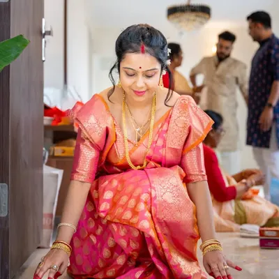 A moment of reverence during a Gruha Pravesh ceremony. The woman, dressed in a stunning pink silk saree, performs a ritual at the threshold of her new home, which is decorated with flowers. This image captures the tradition and grace of the housewarming.