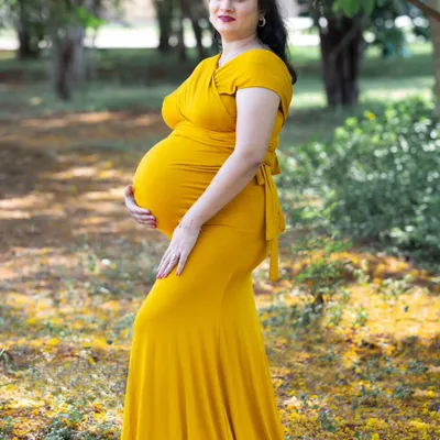 A classic full-length maternity portrait taken outdoors in a natural, leafy environment. The choice of a bright, flowing gown and a delicate floral headdress adds a touch of elegance and celebration to the pregnancy photoshoot.