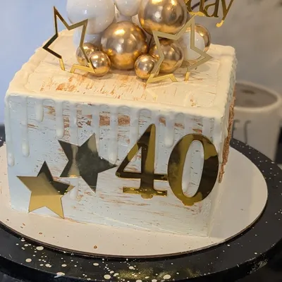 A "Dirty Forty" birthday cake with a touch of glamour. This square cake features a white drip, gold accents, and a cascade of white and gold chocolate balls.