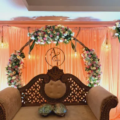 A classic engagement stage design featuring an ornate, carved wooden sofa for the couple. The backdrop is a soft peach drape with a beautiful floral arch and a custom monogram of the couple's initials, all lit by warm, hanging lights.