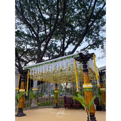 A majestic South Indian wedding mandap at Shankaraa Foundation, featuring dark wooden pillars and a canopy of white chrysanthemums. The hanging temple bells and plantain leaves add layers of authentic tradition.