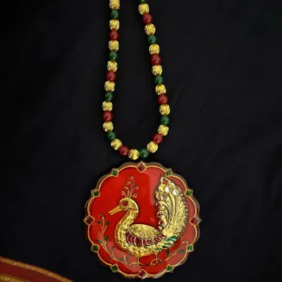 A stunning Tanjore art pendant featuring a peacock motif, crafted with authentic gold foil on a deep red base. The piece is finished with a protective resin coating, ensuring its lasting beauty and shine.