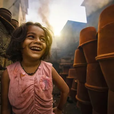 A young girl's infectious laugh lights up a narrow lane filled with terracotta pots. Her expression captures the bright spirit that thrives within the hardworking community of Dharavi.