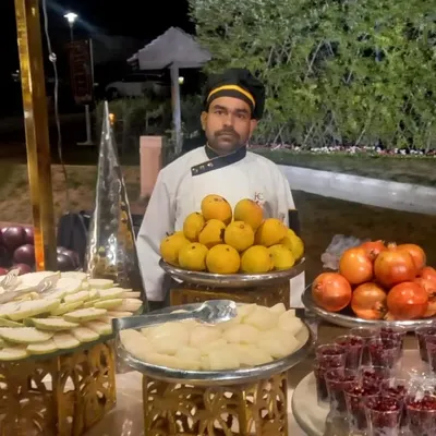 One of my professional chefs standing proudly behind a fresh fruit counter. He is ready to serve guests with a selection of seasonal fruits like mangoes and pomegranates.