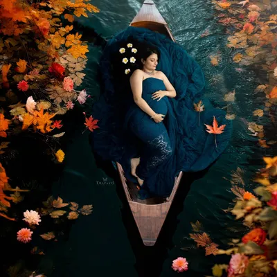A breathtaking and artistic overhead shot. The mother-to-be floats peacefully in a boat, surrounded by autumn leaves, creating a stunning, painterly image of serenity.