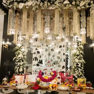 Crafting divine beauty for a Ganpati celebration. This setup features a backdrop of a mirrored floral-patterned panel, framed by pillars of white flowers and topped with a canopy of hanging jasmine and bells.