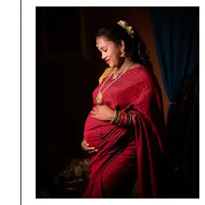 This portrait uses dramatic, low-key lighting to create a sense of intimacy and reverence, reminiscent of a classic painting. The focus is on the mother's gentle expression as she cradles her bump, adorned in a beautiful red silk saree and traditional jewelry.