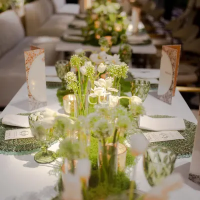 A long-table view of our green and white floral decor concept. The arrangement of fresh flowers, moss placemats, and elegant glassware creates a cohesive and breathtaking dining landscape for a large social gathering.