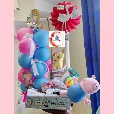 A cheerful baby arrival hamper with a gender-neutral color scheme. It features a Peppa Pig plush, balloons, baby clothes, and toys, all creatively arranged.