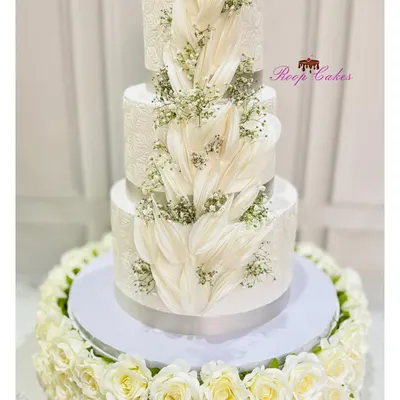 A truly grand statement for a wedding or engagement. This magnificent three-tier cake features intricate white-on-white stenciling, a cascade of wafer paper details, and delicate baby's breath, all surrounded by a bed of fresh white roses.