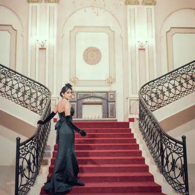A grand, cinematic shot of a woman in an elegant black gown on a red-carpeted staircase. This image evokes the glamour and drama of old Hollywood, perfect for a vintage-themed shoot.