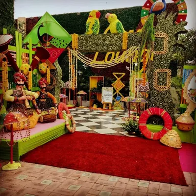 A full-scale Teej festival setup, rich with Rajasthani cultural elements. This photo corner features kathputli puppets, colorful kites, floral peacocks, and a checkered floor, creating a vibrant and traditional celebratory vibe.