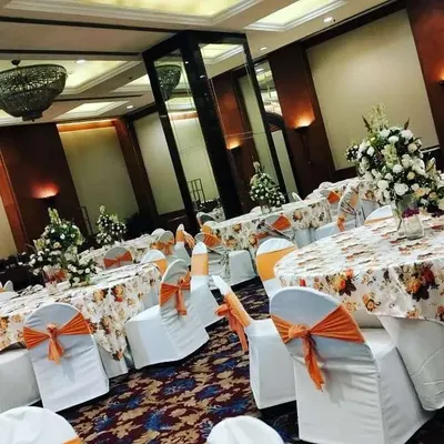 This is a full view of a banquet hall I decorated for a day function. The setup includes round tables with printed cloths, orange bows on the chairs, and fresh floral centerpieces on each table.