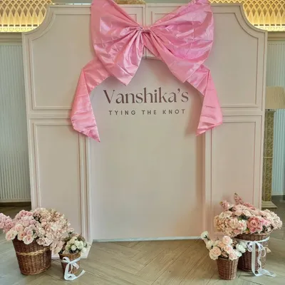 A chic backdrop for "Vanshika's Tying the Knot" bridal shower. The design features a large pink bow on a paneled wall, with baskets of pink roses completing the feminine and celebratory theme.