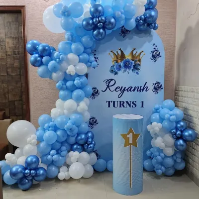 A royal prince theme for a first birthday. This beautiful setup features a custom blue backdrop with a crown motif, an organic balloon arch in shades of blue and white, and a themed pedestal.
