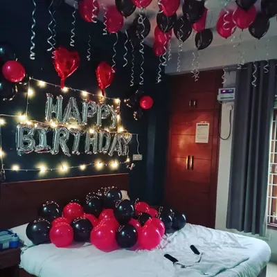 A classic birthday bedroom surprise. The setup includes black and red balloons on the bed and ceiling, with 'Happy Birthday' foil letters illuminated by warm fairy lights on the wall.