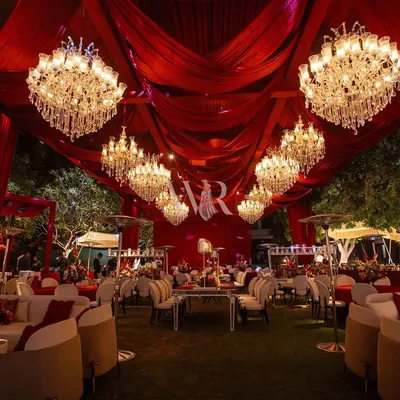 This is the essence of a royal darbar fantasy. The entire venue is enveloped in deep crimson drapes, with a ceiling full of chandeliers casting a golden glow on the plush, elegant seating arrangements below.