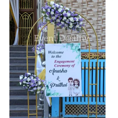 A charming welcome arch for an engagement ceremony. We used a simple golden arch decorated with purple and white floral bunches and a personalized welcome sign featuring a cute caricature of the couple.