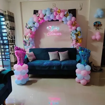 This is a great example of a compact baby shower setup perfect for a living room. It features a balloon arch over the sofa, a "Baby Shower" neon sign, and fun foil balloons shaped like baby feet to complete the look.