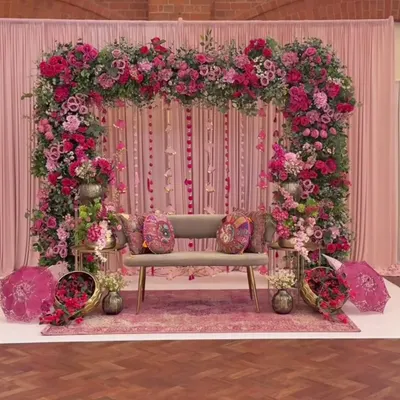 A luxurious Mehendi or engagement stage completely decorated in shades of pink. The setup features a massive floral arch, a backdrop of pink drapes, and elegant seating, creating a romantic and opulent look.
