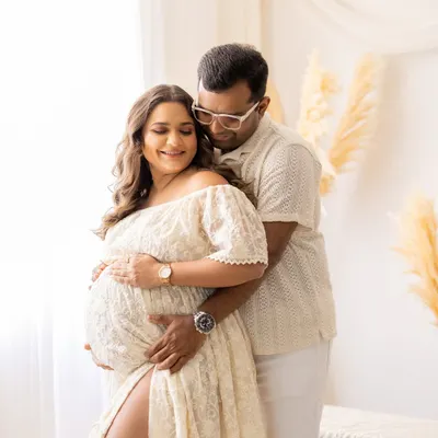 This couple is bathed in soft window light in my studio. The mother-to-be, wearing a beautiful off-white lace gown, cradles her bump while her partner embraces her from behind, creating a tender and romantic maternity portrait.