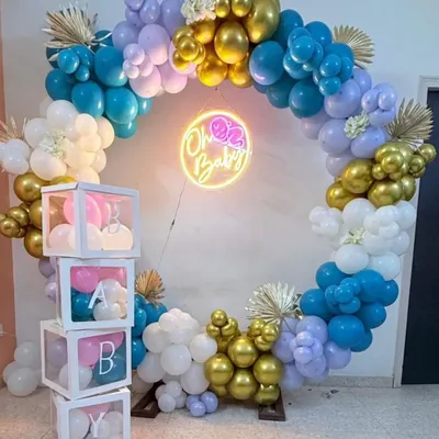 A modern baby shower backdrop featuring a full balloon ring in shades of blue, purple, and gold. The "Oh Baby" neon sign adds a warm glow, and the stacked 'BABY' blocks complete this popular party setup.