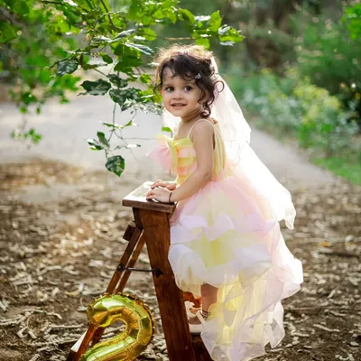 Celebrating turning two! This little girl looks like a fairy in her beautiful pastel dress, posing on a ladder with a "2" balloon in a magical, sunlit forest setting.