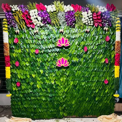 A lush green backdrop made entirely of leaves, decorated with pink lotus cutouts. The top is adorned with a mix of vibrant fresh flowers, creating a natural and divine look for a baby shower.