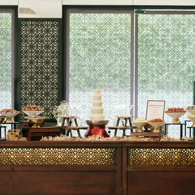 An elaborate dessert buffet featuring a white chocolate fountain and an array of Indian sweets. Our buffet setups are designed to be a visual spectacle, inviting guests to indulge in a thoughtfully curated selection of treats.