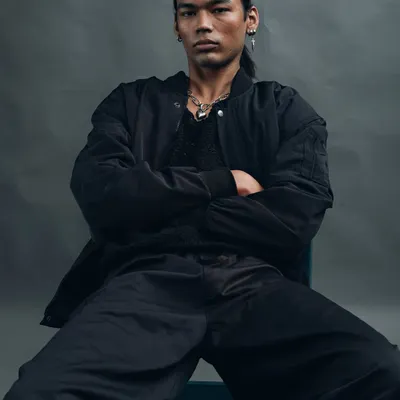 A powerful, seated portrait with a moody, grey background. The model's crossed arms and direct gaze convey a sense of strength and confidence. The lighting is soft yet dramatic, sculpting his features.