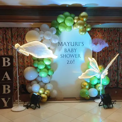 A modern and minimalist baby shower setup with a green and gold balloon garland on a white arched backdrop. The look is completed with illuminated butterfly props and wooden 'BABY' blocks.