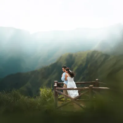 A couple shares an intimate moment on a wooden viewpoint, overlooking a breathtaking valley of green mountains. The soft mist adds a dreamy quality to this stunning landscape portrait.