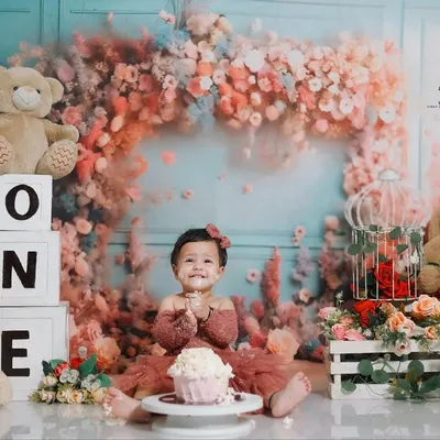A perfect first birthday celebration. This little girl is all smiles, ready to dig into her cake in a stunning studio setup featuring a floral arch, teddy bears, and 'ONE' blocks.