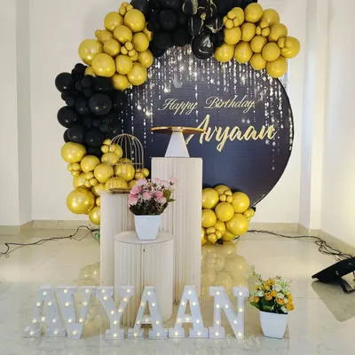 A chic and modern birthday setup with a black and gold balloon arch. This decoration includes a personalized backdrop with a starlight effect, elegant white plinths, and marquee letters for the name.
