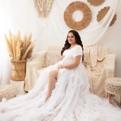A beautiful full-length portrait in the boho setup. The mother-to-be is seated on the couch, with the long, ruffled train of the white gown cascading around her.