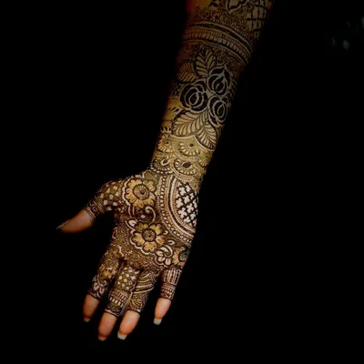A stunning, professional shot of a full-arm bridal mehendi stain. The dark, rich color and intricate floral details are captured beautifully against the black background.