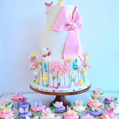 This first birthday dessert spread features a three-tiered floral cake with a large fondant bow, surrounded by matching cupcakes with sugar flowers and butterfly toppers.
