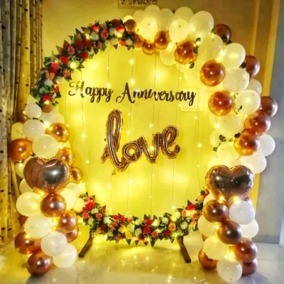 A romantic anniversary decoration for a home celebration. This setup features a circular backdrop with white and gold balloons, flowers, fairy lights, and a 'love' foil balloon.
