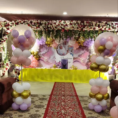 This is a lavish stage setup for a naming ceremony or first birthday. It includes a ceiling covered in artificial flowers, a custom backdrop, and elegant balloon pillars in pastel and gold tones.