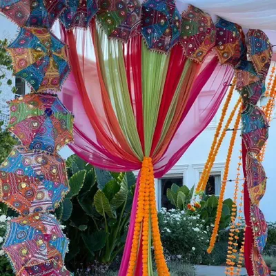 An outdoor garden setup for a Mehendi, featuring a canopy of colorful drapes and Rajasthani umbrellas. The decor is tied together with bright orange marigold garlands.