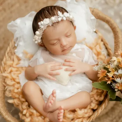 A peaceful newborn girl, dressed in a delicate white outfit and floral headband, sleeping soundly in a cozy woven basket. We focus on creating a serene atmosphere for these early sessions.